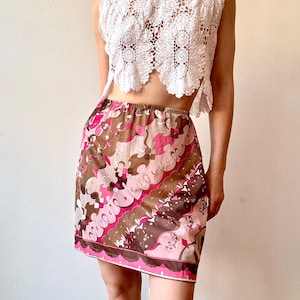 Può includere: Un top all'uncinetto bianco a trama aperta abbinato a una minigonna. La gonna presenta un motivo colorato con vortici e motivi floreali rosa, marroni e bianchi. La gonna ha un bordo rosa.