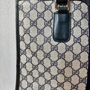 May include: A vintage handbag with a repeating pattern of interlocking Gs in navy blue on a beige background. The bag has navy blue leather trim, a navy blue handle, and gold-colored hardware. A navy blue and red stripe is on the side.