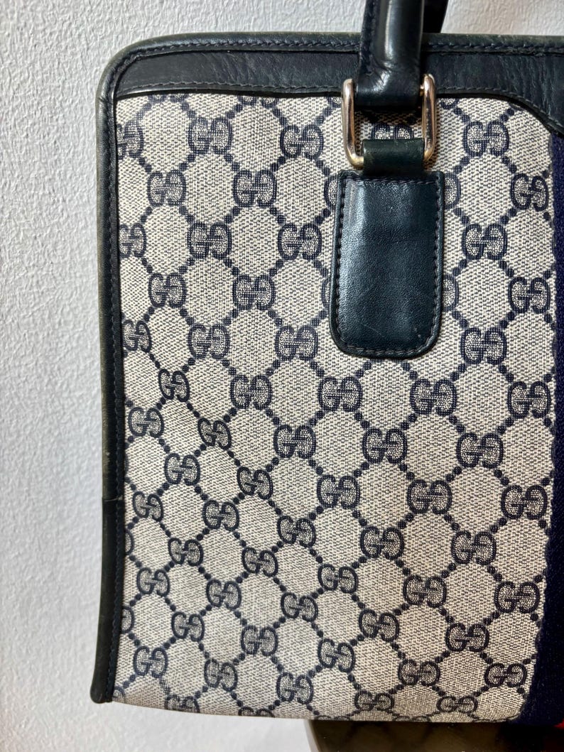 May include: A vintage handbag with a repeating pattern of interlocking Gs in navy blue on a beige background. The bag has navy blue leather trim, a navy blue handle, and gold-colored hardware. A navy blue and red stripe is on the side.