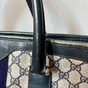 May include: Close-up of a handbag with a black leather handle and trim. The bag features a beige canvas with a repeating dark blue interlocking "G" pattern. A vertical blue stripe is visible on the side.