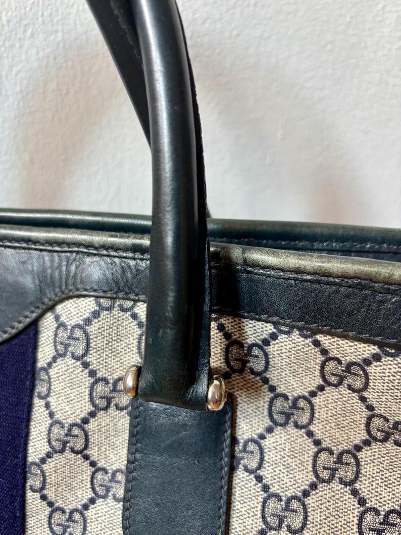 May include: Close-up of a handbag with a black leather handle and trim. The bag features a beige canvas with a repeating dark blue interlocking "G" pattern. A vertical blue stripe is visible on the side.