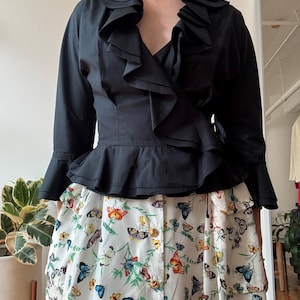 May include: A black ruffled blouse with a wrap style and long sleeves. The blouse has a deep V-neckline and a gold chain necklace. The model is wearing a white skirt with a butterfly print.