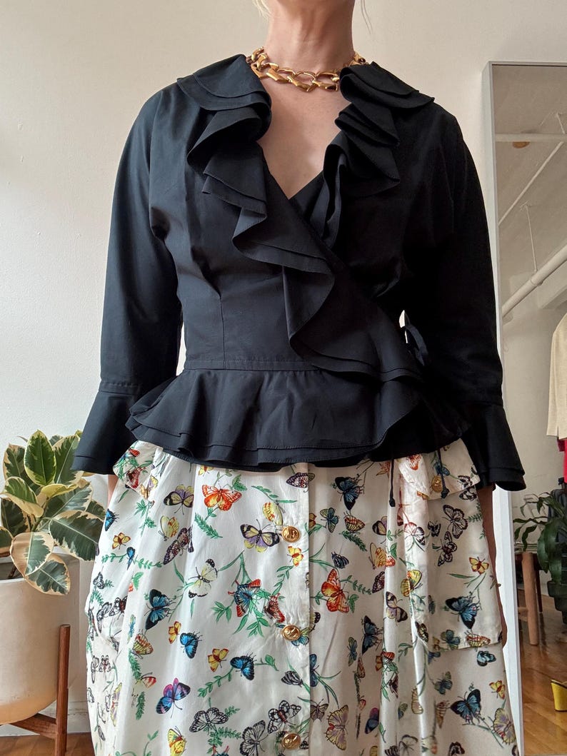 May include: A black ruffled blouse with a wrap style and long sleeves. The blouse has a deep V-neckline and a gold chain necklace. The model is wearing a white skirt with a butterfly print.