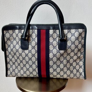 May include: A vintage handbag with a repeating pattern of interlocking "G" logos in beige and navy blue. The bag features a central vertical stripe in navy blue and red. It has black leather handles and trim.