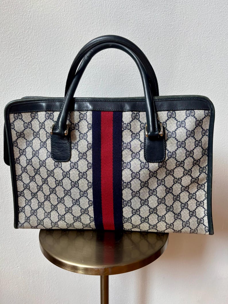 May include: A vintage handbag with a repeating pattern of interlocking "G" logos in beige and navy blue. The bag features a central vertical stripe in navy blue and red. It has black leather handles and trim.