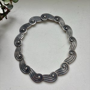 May include: A silver-toned necklace composed of repeating, interconnected links. Each link features a stylised, swirling wave pattern with a small, raised circular bead at the centre of the spiral. The metal has a polished finish with darkened recessed areas to accentuate the curved, flowing design of the individual segments.