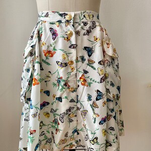 May include: A white skirt with a butterfly print and gold buttons. The skirt has two large pockets on the sides.