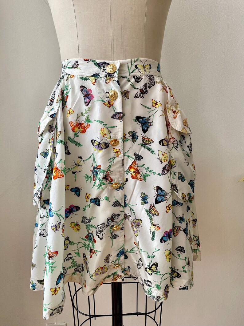 May include: A white skirt with a butterfly print and gold buttons. The skirt has two large pockets on the sides.