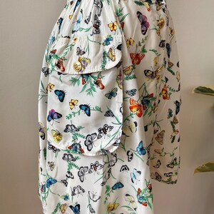 May include: A white skirt with a colorful butterfly print. The skirt has a large patch pocket on the left side and gold buttons on the waistband.