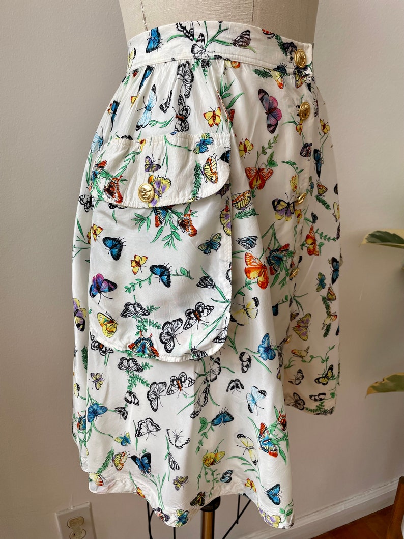 May include: A white skirt with a colorful butterfly print. The skirt has a large patch pocket on the left side and gold buttons on the waistband.