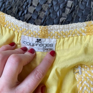 May include: A yellow and white tweed jacket with a Courr&egrave;ges Paris label. The jacket has a white lining and a metal hook closure.