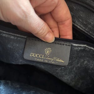 May include: Close-up of a black leather bag interior. A rectangular label with gold lettering reads "GUCCI Luxury Collection MADE IN ITALY". The bag's interior shows a textured, worn appearance.