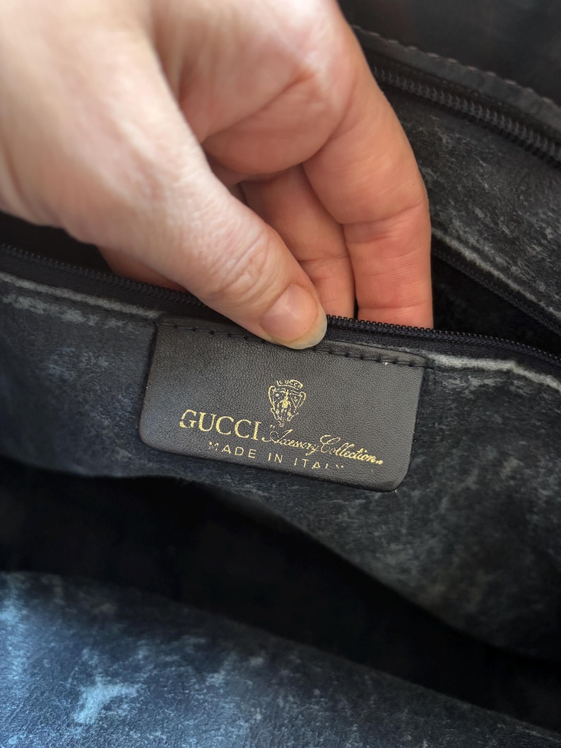 May include: Close-up of a black leather bag interior. A rectangular label with gold lettering reads "GUCCI Luxury Collection MADE IN ITALY". The bag's interior shows a textured, worn appearance.