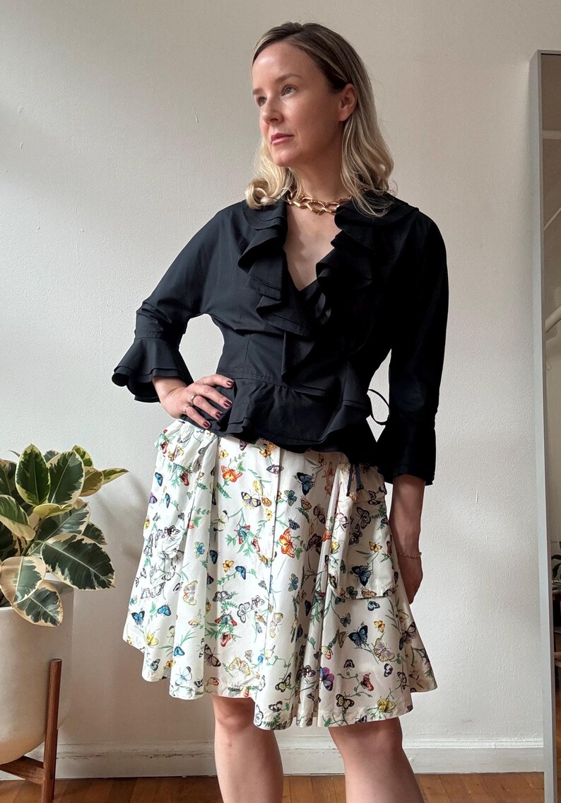 May include: A woman wearing a black ruffled blouse with a gold chain necklace and a white skirt with a butterfly print.