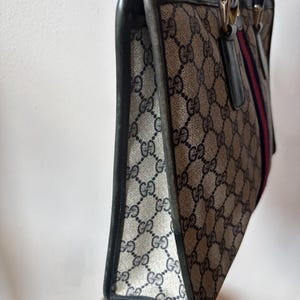 May include: A vintage handbag with a repeating GG logo pattern in beige and navy. The bag features black leather trim, handles, and a red and navy stripe detail. The bag is a classic tote style.