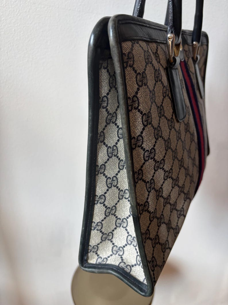 May include: A vintage handbag with a repeating GG logo pattern in beige and navy. The bag features black leather trim, handles, and a red and navy stripe detail. The bag is a classic tote style.