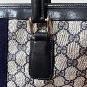 May include: Close-up of a tote bag with a repeating pattern of interlocking Gs in a light gray and beige color. The bag has a navy blue stripe and black leather handles with silver-tone hardware.