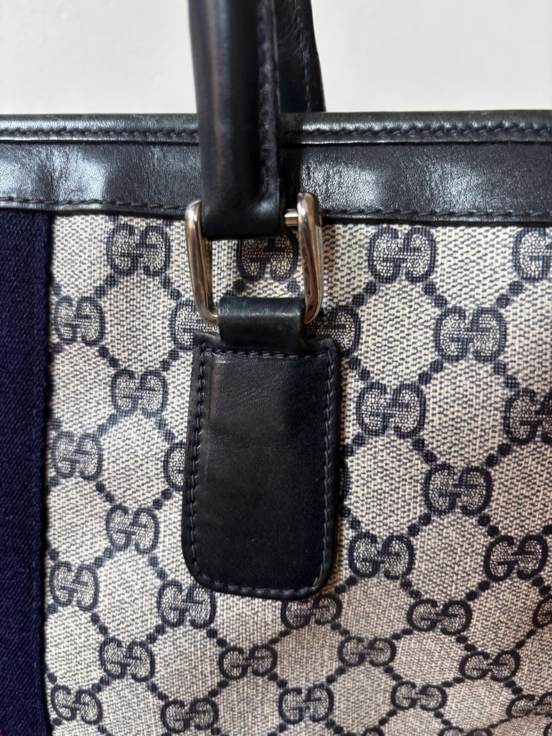 May include: Close-up of a tote bag with a repeating pattern of interlocking Gs in a light gray and beige color. The bag has a navy blue stripe and black leather handles with silver-tone hardware.