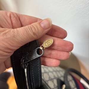 May include: Close-up of a black leather handbag zipper with a gold-toned pull featuring a double-G logo. The bag has a beige and gray pattern with a red and blue stripe.