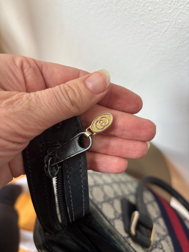 May include: Close-up of a black leather handbag zipper with a gold-toned pull featuring a double-G logo. The bag has a beige and gray pattern with a red and blue stripe.