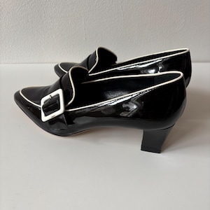 Puede incluir: Par de mocasines de tacón de charol negro con ribete blanco y hebilla blanca. Los zapatos tienen una punta puntiaguda y un tacón de bloque pequeño. Los mocasines se exhiben sobre una superficie blanca.