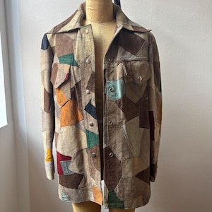 May include: A long-sleeve jacket constructed from multi-colored suede leather patches in shades of brown, tan, green, yellow, and red. The garment features a pointed collar, two chest pockets with flaps, and a front button closure with metal snaps.