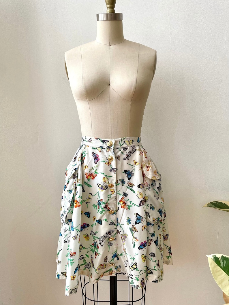 May include: A white skirt with a butterfly print. The skirt has a button closure and two pockets on the sides. The skirt is on a white mannequin.