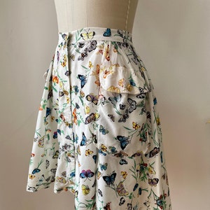 May include: A white skirt with a butterfly print. The skirt has a ruffle at the bottom and a pocket on the side.