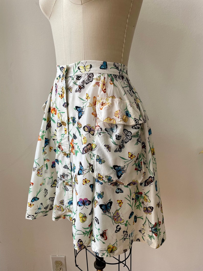 May include: A white skirt with a butterfly print. The skirt has a ruffle at the bottom and a pocket on the side.