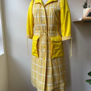 May include: A yellow and white plaid coat with a yellow solid color lining. The coat has a drawstring waist and two large pockets.