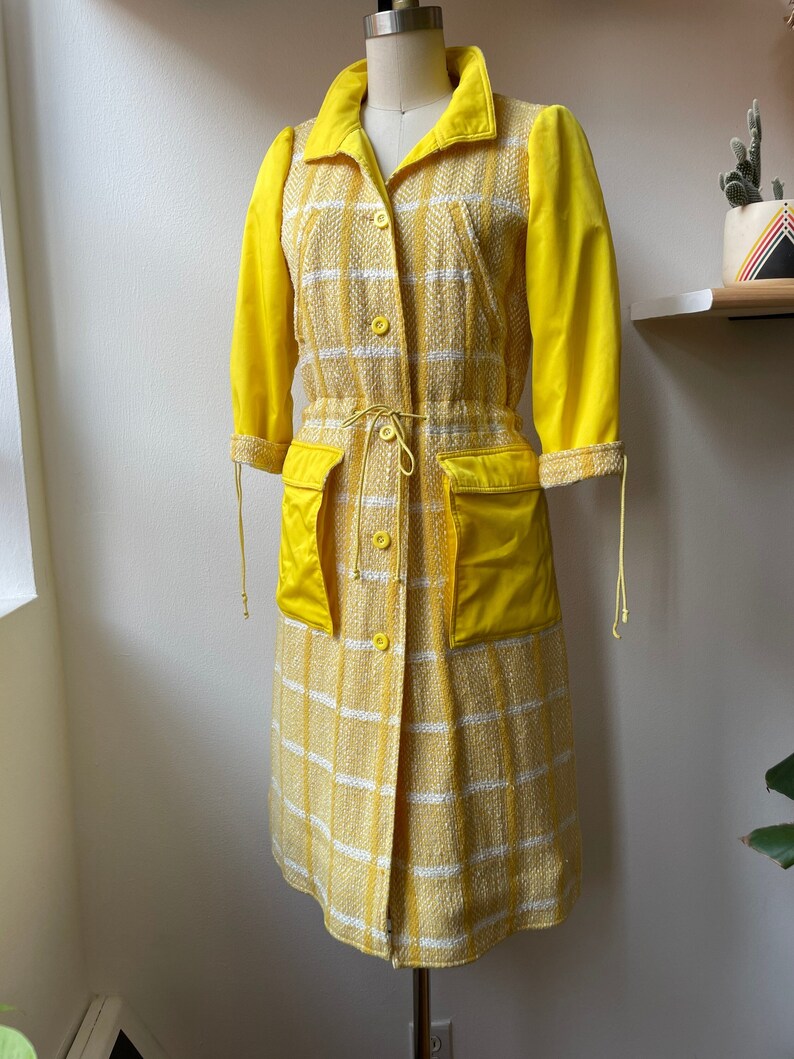 May include: A yellow and white plaid coat with a yellow solid color lining. The coat has a drawstring waist and two large pockets.