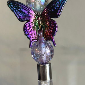 Puede incluir: Una corona plateada con pedrería iridiscente se asienta sobre una cuenta de vidrio transparente con purpurina iridiscente. Un amuleto de mariposa iridiscente morado y verde está unido a la cuenta. Debajo del amuleto de mariposa hay una cuenta de vidrio morado con purpurina iridiscente. La parte inferior del amuleto es un tubo de vidrio transparente lleno de cuentas iridiscentes.