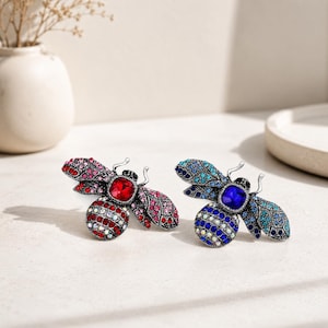 May include: Two bee-shaped brooches crafted from silver-toned metal. One brooch features red and pink crystals with a central red stone, while the other displays blue and turquoise crystals with a central deep blue stone. Both have detailed wings.