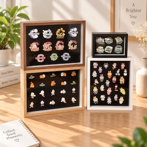 May include: Three rectangular display cases with wooden frames in dark brown, light brown, and white, featuring black interiors filled with various small enamel pins. The pins depict diverse characters, logos, and text, organized in grid-like arrangements.
