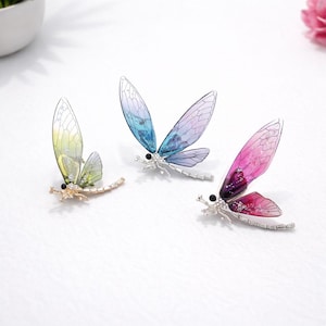 May include: Three dragonfly brooches with translucent wings in shades of green, blue, and pink. The dragonfly bodies are silver and gold-toned, with black eyes and crystal accents. These decorative brooches are perfect for adding a touch of nature-inspired elegance.