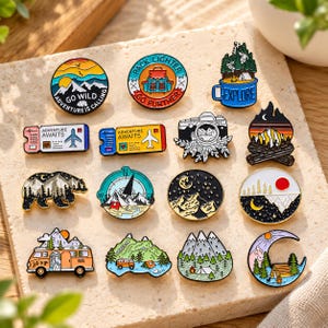 May include: A collection of fourteen enamel pins featuring outdoor and travel themes. Designs include mountains, a camera, a camper van, a compass, a bear, a crescent moon, and ticket stubs with the text Adventure Awaits, Go Wild, and Explore.
