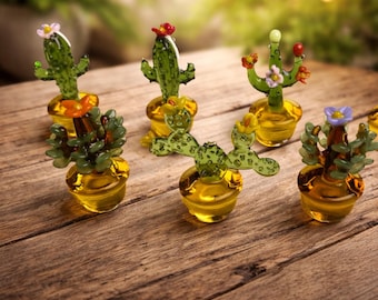 May include: A collection of miniature glass cactus sculptures. Each cactus is a different shape and color, with details like flowers and spines. They are set in small, yellow-tinted glass pots, arranged on a wooden surface.