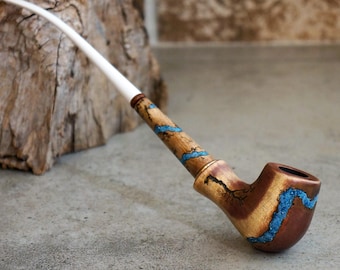 May include: A handcrafted tobacco pipe with a brown bowl and a light-colored stem. The wooden stem is decorated with blue accents. The pipe rests on a gray surface, with a piece of wood in the background.