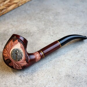 May include: A brown wooden smoking pipe with a carved design of a bird on the bowl. The pipe has a black stem with a gold band.