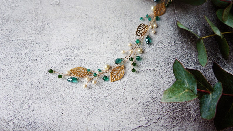Puede incluir: Un peine de pelo de hoja dorada y verde con perlas y cuentas de color verde esmeralda. El peine est&aacute; sobre un fondo gris texturizado con hojas verdes.