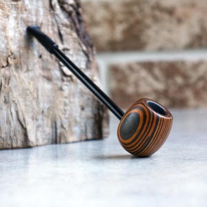 May include: A wooden pipe with a black stem and a bowl featuring a striped pattern in shades of brown and black. The pipe is angled against a piece of wood, with a brick wall in the background.