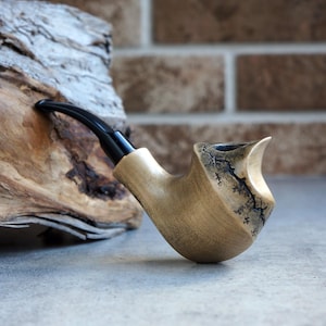 May include: A handcrafted wooden pipe with a curved black stem. The pipe has a light-colored bowl with dark, intricate patterns near the rim. The pipe rests on a gray surface, with a piece of driftwood in the background.