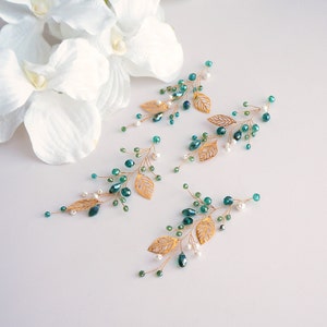Puede incluir: Cuatro pinzas para el pelo de hoja de oro con cuentas verde azulado y perlas blancas. Las pinzas est&aacute;n dispuestas sobre una superficie blanca.