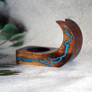 May include: A wooden ring holder with a crescent moon shape. The holder is made of dark wood with a blue resin inlay that resembles a river.
