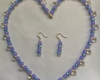 May include: A delicate necklace and earrings set featuring light purple and clear glass beads. The necklace is a long strand of beads with a heart shape formed by the beads.
