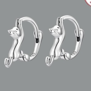 May include: A pair of silver hoop earrings with a small, detailed cat design. The cat is hanging from the hoop and appears to be climbing. The earrings are made of sterling silver.