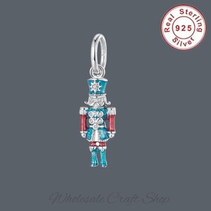 May include: A silver charm depicting a toy soldier, with blue, red, and white enamel detailing. The charm has a loop for attaching to jewellery. A red circle with the text "Real Sterling 925 Silver" is visible.