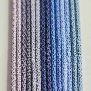 May include: Close-up of several strands of cord in a gradient of blues and purples. The cords are tightly woven, creating a textured surface. The color transitions from light lavender to deep indigo.
