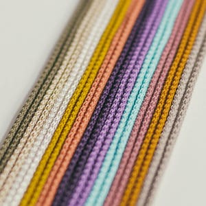 May include: Close-up of a collection of colorful cords in various shades, including beige, yellow, purple, and blue. The cords are tightly woven and arranged in parallel lines, creating a textured pattern. The cords are likely used for crafting or jewelry making.