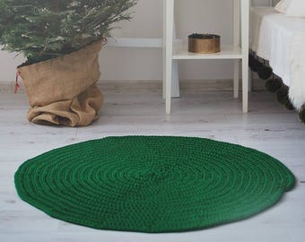 May include: A green crocheted round rug with a white wooden side table with a drawer and a shelf. The table has a white wooden sign that says "Selima" on it. There is a small potted Christmas tree on the table.